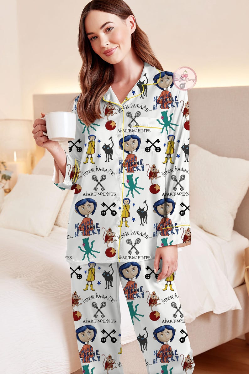 May include: White pyjama set with a repeating pattern of cartoon characters, cats, keys, and the text "PINK PALACE" and "BE BRAVE BE TRICKY". The pyjamas have yellow trim and are worn by a person holding a white mug.