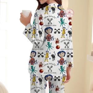 May include: White pyjama set with a repeating pattern of cartoon characters, black cats, and text. The pyjama top and trousers feature a whimsical design with yellow accents and the words "PINK PALACE" and "APARTMENTS".
