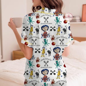 May include: White pyjama set with a colourful pattern featuring cartoon characters, keys, and the text "PINK PALACE" and "BE TRICKY". The short-sleeved top and shorts have yellow trim.