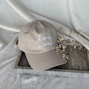 Peut inclure: Casquette de baseball beige avec une visi&egrave;re incurv&eacute;e. Le texte "dog mom" est brod&eacute; en blanc sur le devant. La casquette est pos&eacute;e sur un plateau gris avec une branche de fleurs blanches.