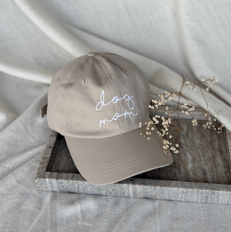 Peut inclure: Casquette de baseball beige avec une visi&egrave;re incurv&eacute;e. Le texte "dog mom" est brod&eacute; en blanc sur le devant. La casquette est pos&eacute;e sur un plateau gris avec une branche de fleurs blanches.