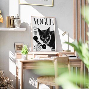May include: A framed Vogue magazine cover featuring a black cat wearing a pearl necklace and holding a vintage telephone. The cover includes the Vogue logo and the words "I AM STYLE ICON HOLY CHIC LOOK". The frame is light brown, and the image is displayed on a wooden desk.