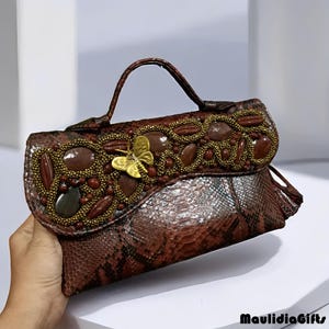 May include: Brown snakeskin handbag with a handle and flap closure. The flap is embellished with brown stones, gold beads, and a gold butterfly. The bag has a small handle and a tassel detail.