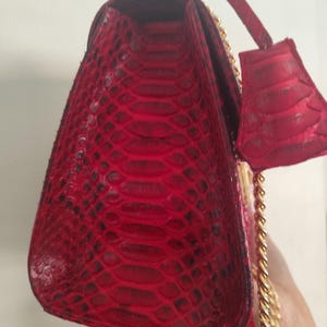 Puede incluir: Un bolso de mano rojo vibrante con un patr&oacute;n de piel de serpiente texturizado en el exterior. El bolso incluye un asa superior estructurada, una etiqueta de equipaje a juego en piel de serpiente roja y una correa de cadena dorada.
