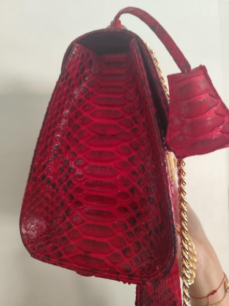 Puede incluir: Un bolso de mano rojo vibrante con un patr&oacute;n de piel de serpiente texturizado en el exterior. El bolso incluye un asa superior estructurada, una etiqueta de equipaje a juego en piel de serpiente roja y una correa de cadena dorada.