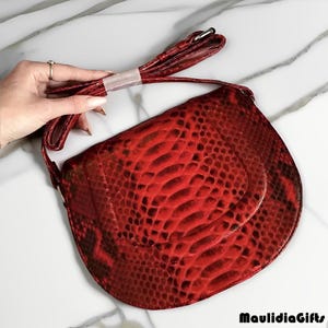 May include: A red, snake-skin patterned crossbody bag with a matching strap. The bag has a curved shape and a flap closure. The strap is held by a hand with a ring. The bag is on a white marble surface.
