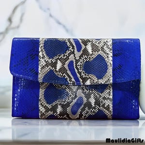 May include: A rectangular clutch with a flap closure. The clutch features a vibrant blue leather body with a central panel of snakeskin in shades of gray, black, and white. The brand name "MaulidiaGifts" is visible in the bottom right corner.