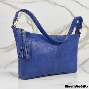 May include: A blue handbag with a textured snakeskin pattern. The bag has a shoulder strap, a zipper closure, and a decorative tassel. The bag is approximately 12 inches wide and 8 inches tall.