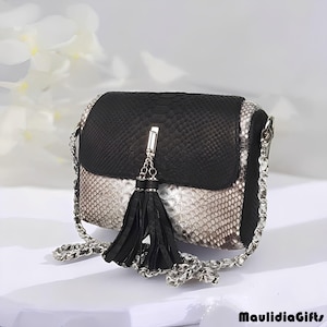 May include: A black and white snakeskin-patterned crossbody bag with a silver chain strap. The bag features a black flap and a black tassel. The bag is a small, rectangular shape.