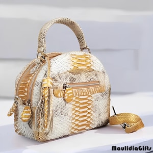 May include: A white, gold, and gray snakeskin handbag with a top handle and a matching small belt. The bag has a rounded shape with a zipper closure and a decorative tassel. The bag is sitting on a white surface.