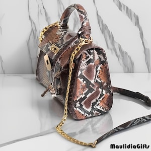 May include: A brown and tan snakeskin-patterned handbag with a gold chain strap and handle. The bag has a rectangular shape with a flap closure and a gold clasp. The bag also has a detachable snakeskin strap. The brand name "MaulidiaGifts" is printed in black.