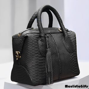 May include: Black handbag with a textured snakeskin pattern. The bag has two curved handles, a decorative tassel, and gold-colored hardware. The bag is sitting on a white surface.