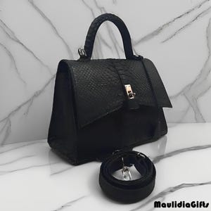 May include: A black handbag with a textured snakeskin pattern. The bag has a top handle and a detachable shoulder strap. A gold-colored clasp secures the flap closure. The bag is sitting on a white marble surface.