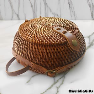 May include: A round, woven rattan backpack with a brown leather strap and clasp. The bag has a circular lid and a leather strap closure. The bag is a light brown color and has a natural, textured appearance.