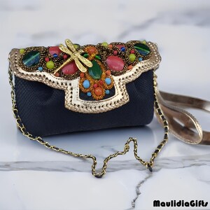 May include: A small, black handbag with a gold chain strap. The flap is adorned with colorful gemstones, beads, and a gold dragonfly. The edges are trimmed with gold, and the bag has a gold chain strap.