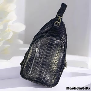 May include: Black leather sling bag with a reptile skin texture. The bag features a zippered main compartment and a smaller front pocket with a silver zipper. It has a single adjustable strap and a metal clasp. The bag is designed to be worn across the body.
