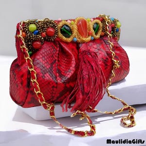 May include: A red snakeskin clutch with a gold chain strap. The top of the bag is adorned with colorful beads and gemstones. A red fringe hangs from the front of the bag.