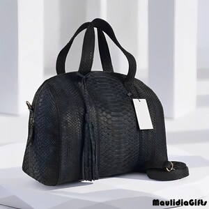 May include: A black leather handbag with a snakeskin texture. The bag has two handles, a detachable shoulder strap, and a tassel. A blank white tag hangs from the side. The bag is on a white surface.