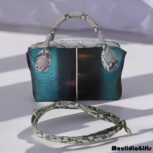 May include: A teal and black handbag with a snakeskin pattern handle and trim, accompanied by a matching snakeskin bracelet. The bag has a structured shape and a contrasting vertical panel. The bracelet is curved and lies in front of the bag.
