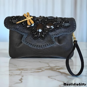 May include: A black clutch with a textured pattern, embellished with black beads and stones. A gold dragonfly charm accents the flap. The clutch has a wrist strap and a gold-tone chain.