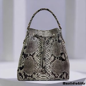May include: A bucket-style handbag with a snakeskin pattern in shades of gray, black, and white. The bag has a drawstring closure and a top handle. Gold-colored hardware accents the handle attachments. The bag is sitting on a white surface.
