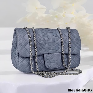 May include: A blue-grey handbag with a chain strap. The bag has a quilted pattern and a flap closure. The chain strap is silver. The bag is made of a textured material, possibly leather or a leather-like material. The bag is sitting on a white surface.