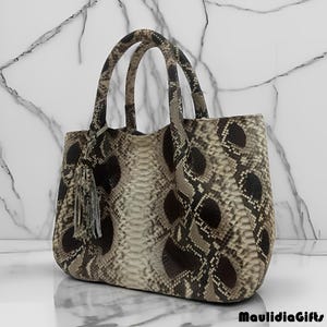 May include: A snakeskin-patterned tote bag with two rounded handles and a tassel. The bag features a mix of brown, black, and cream tones, with a textured surface. The bag is designed for carrying items.