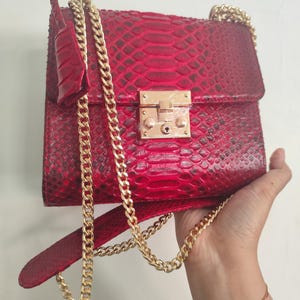Puede incluir: Un bolso de mano rojo vibrante con un patr&oacute;n de piel de serpiente texturizado, un cierre de solapa rectangular y un broche de metal dorado brillante. El bolso incluye una correa larga de cadena dorada y una hombrera de piel a juego.