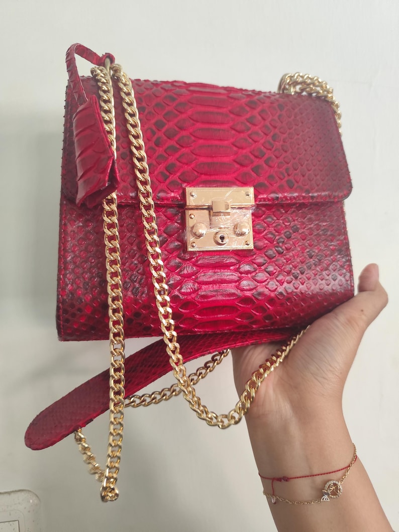 Puede incluir: Un bolso de mano rojo vibrante con un patr&oacute;n de piel de serpiente texturizado, un cierre de solapa rectangular y un broche de metal dorado brillante. El bolso incluye una correa larga de cadena dorada y una hombrera de piel a juego.