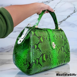 May include: A bright green handbag with a snakeskin pattern. The bag has a top handle, a clasp closure, and gold-colored hardware. The bag is held up by a person's hand.