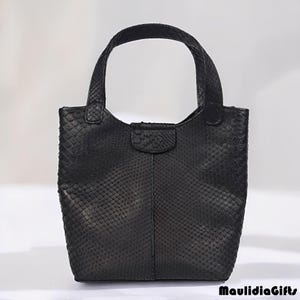 May include: A black handbag with a textured, snake-like pattern. The bag has a rectangular body with a rounded top and a single handle. The handle is attached to the bag with small, reinforced loops. The bag is made of a material that resembles snakeskin.
