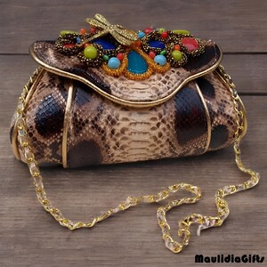 May include: A snakeskin-patterned handbag with a gold chain strap. The flap is adorned with colorful beads and a gold dragonfly. The bag has a gold trim and a decorative clasp.