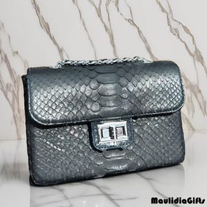 May include: A gray handbag with a snakeskin pattern. The bag has a silver chain strap and a silver clasp closure. The bag is rectangular and has a flap closure. The brand name "MaulidiaGifts" is printed at the bottom.