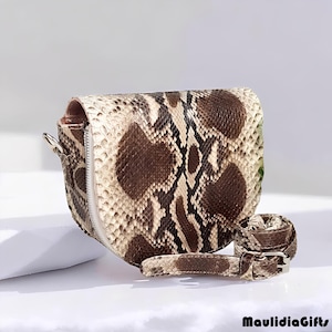 May include: A snakeskin-patterned crossbody bag with a matching belt. The bag features a curved shape, a zipper closure, and a silver-tone metal zipper pull. The belt has a silver-tone buckle. The bag and belt are in shades of brown, tan, and cream.