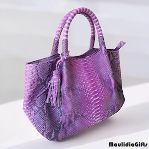 May include: A purple handbag with a snakeskin pattern. The bag has a rounded shape, a pair of handles with white and purple stripes, and a decorative tassel. The bag is made of a textured material and has a zipper closure.