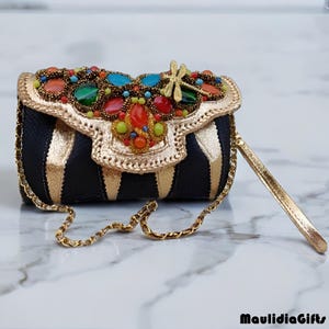 May include: A small, black and gold striped handbag with a gold chain strap and a gold handle. The flap is adorned with colorful gemstones and a gold dragonfly. The bag is embellished with gold trim.