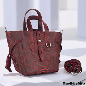 May include: A red and black snakeskin-patterned tote bag with a matching shoulder strap. The bag has two handles and a flap closure with a silver-tone clasp. The strap is rolled up next to the bag. The bag is on a white surface.