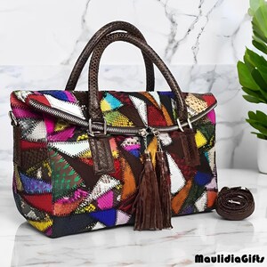 May include: A colorful patchwork leather handbag with a zipper closure and brown handles. The bag features a variety of colors and textures, with a brown tassel and a detachable shoulder strap. The bag is sitting on a white marble surface.