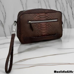 May include: A brown leather clutch with a snakeskin pattern. The rectangular bag has a silver zipper and a wrist strap attached with a silver clasp. The bag is on a white marble surface. The text "MaulidiaGifts" is in the bottom right corner.