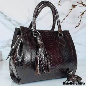 May include: A dark brown, textured handbag with a matching tassel and handle. The bag has a zipper closure and a small, decorative charm. The bag is made of a material that resembles snakeskin. The brand name "MaulidiaGifts" is visible.