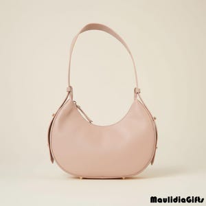 May include: A blush-colored, crescent-shaped handbag with a single shoulder strap. The bag features a zipper closure and small gold-toned studs on the bottom. The bag is made of a smooth, leather-like material.