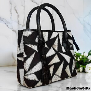 May include: A black and white geometric patterned handbag with black handles and a tassel. The bag features a patchwork design of triangles and other shapes. The bag is sitting on a white surface.