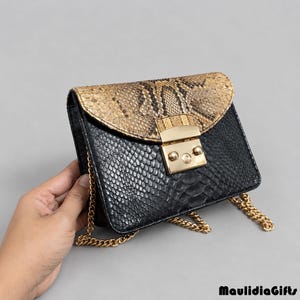 May include: A structured handbag featuring a black snakeskin-textured body and a tan snakeskin-textured flap. The bag includes a gold-toned metal push-lock closure and a gold-toned chain shoulder strap. The text MaulidiaGifts is visible.