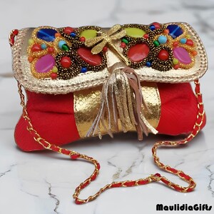 May include: A red and gold clutch with a decorative flap adorned with colorful beads and a gold dragonfly clasp. The clutch features a gold chain strap and a tassel. The brand name "MaulidiaGifts" is visible in the bottom right corner.