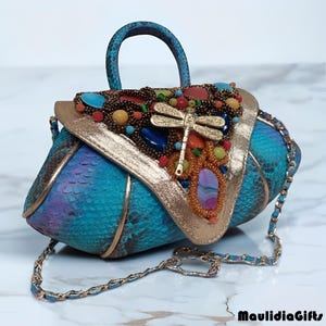May include: A handcrafted, turquoise handbag with a snakeskin pattern. The bag features a gold-colored dragonfly, colorful beads, and a gold-colored trim. It has a matching handle and a silver chain strap.