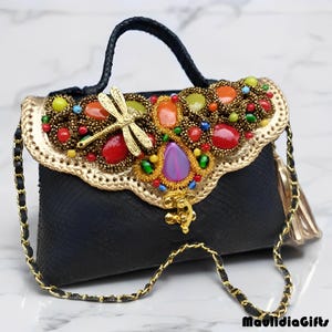May include: Black handbag with a gold chain strap and handle. The flap is adorned with colorful gemstones, beads, and a gold dragonfly. The edges are trimmed with gold, and a gold tassel hangs from the side.