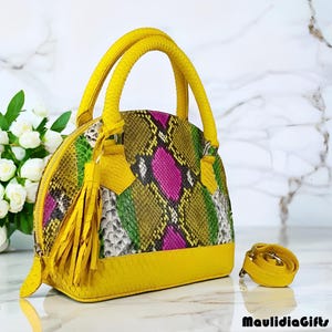 May include: A vibrant, dome-shaped handbag with a snakeskin pattern in shades of green, pink, and yellow. The bag features bright yellow handles, a matching base, and a tassel. An additional yellow strap is included. The bag is a stylish accessory.