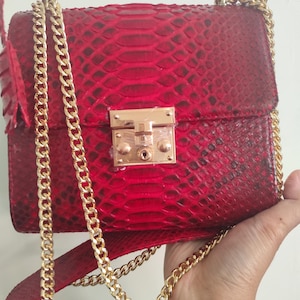 Puede incluir: Un bolso de mano rojo vibrante con un patr&oacute;n de piel de serpiente texturizado. El bolso es rectangular con un cierre de metal dorado en la solapa frontal. Incluye una correa de piel de serpiente roja y una cadena de eslabones de metal dorado.
