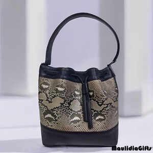 May include: A black and snakeskin-patterned bucket bag with a single handle. The bag features a drawstring closure and a solid black base. The snakeskin pattern includes shades of brown, beige, and white. The bag is designed with a sleek, modern aesthetic.