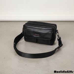 May include: Black leather crossbody bag with a rectangular shape and a long, adjustable strap. The bag features a front flap pocket and a small rectangular accent on the front. The bag is designed for carrying essentials.